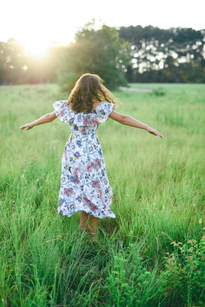 Person standing in a grassy field with arms outstretched at sunset.
